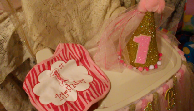 One-year birthday party hat and bib in pink and gold, sitting on a baby's high chair.